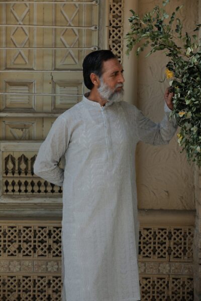 A fashionable man wearing a luxury grayish-white embroidered kurta for men, looking thoughtfully to the side against a neutral heritage backdrop.