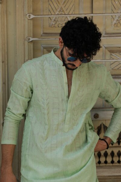 A fashionable man wearing a luxury mint green embroidered kurta for men, leaning against a rustic wall