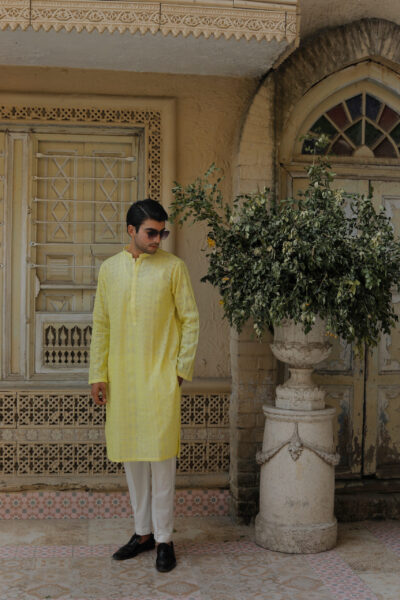 Man wearing a premium lemon yellow embroidered Chikankari kurta with white trousers and sunglasses in a classic architectural setting.