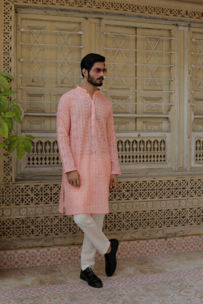 A stylish man with a beard leaning against a carved wooden door, wearing a luxury peach-colored hand-embroidered kurta.