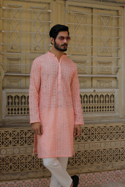 A stylish man with a beard leaning against a carved wooden door, wearing a luxury peach-colored hand-embroidered kurta.